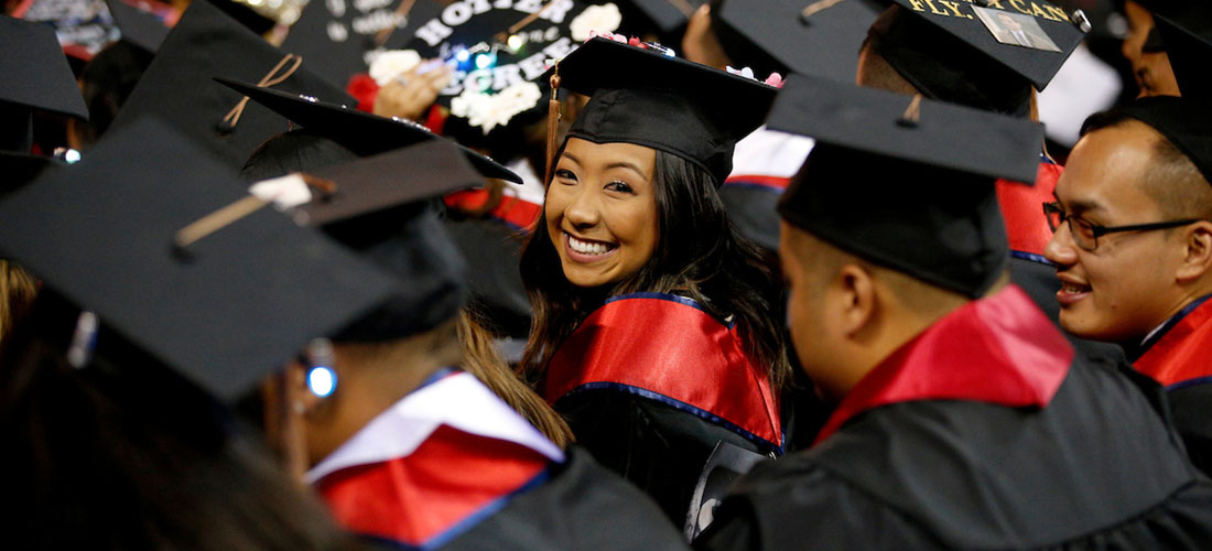 An Asian woman in a cap and gown smiles over her shoulder during commencement